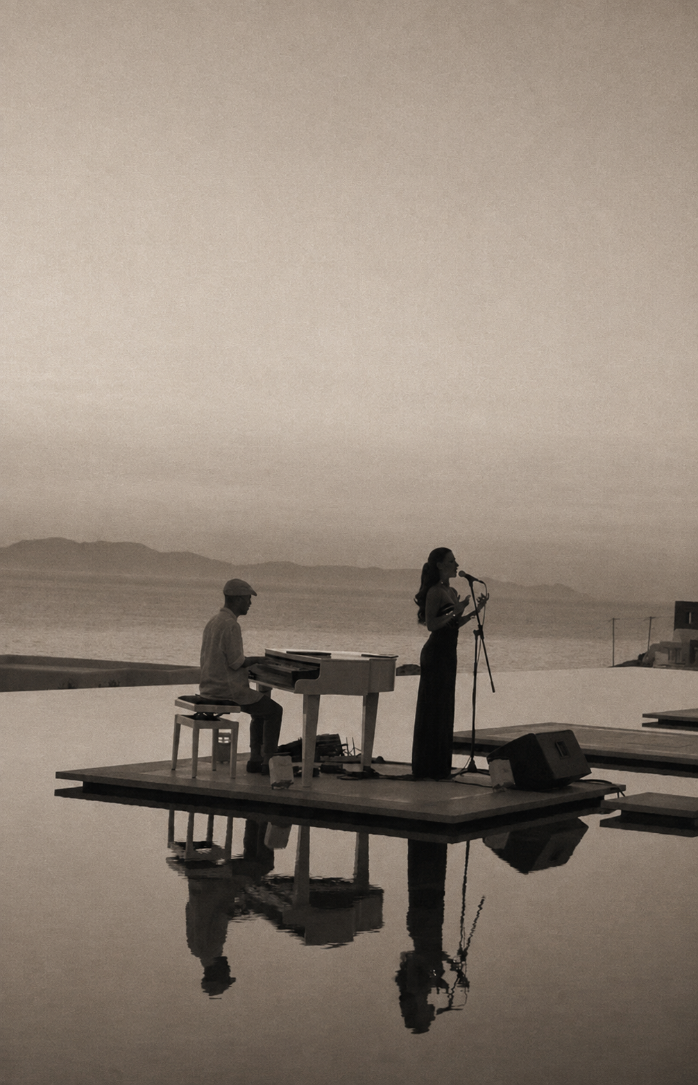 Man plays a piano on a floating stage while a woman sings into a microphone, against a calm sea at sunset (sepia-toned).