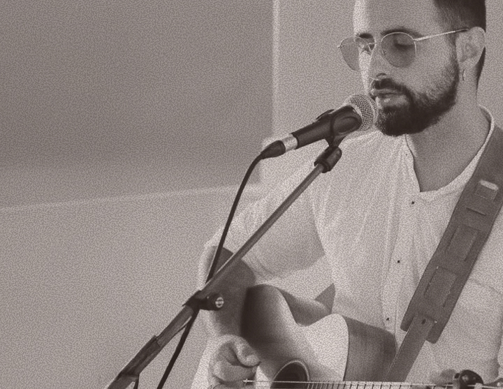 Bearded man wearing glasses sings into a microphone while playing an acoustic guitar, mid-performance.