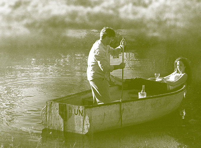 Two people in a small rowboat on a calm river; one man paddles with a pole while the other reclines with a wine glass and bottle nearby, hull marked UN.