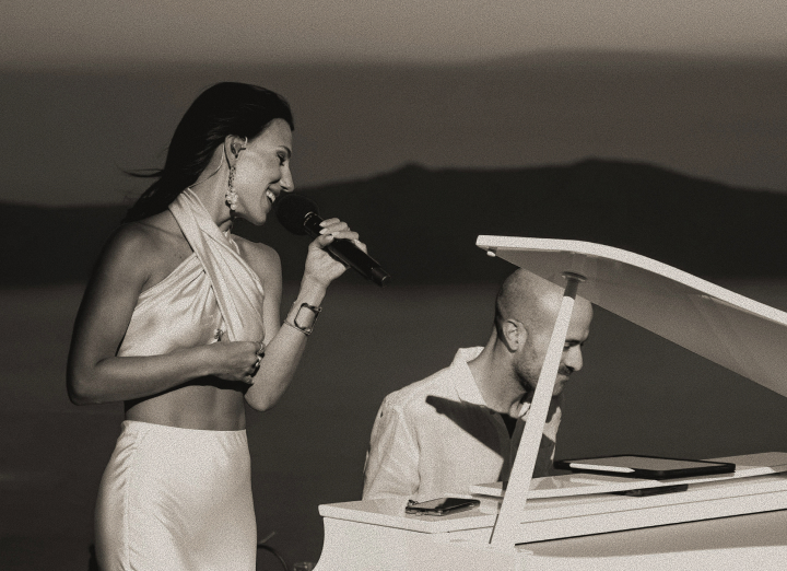 Woman in a white dress sings into a microphone as a man plays a white grand piano on a beach at sunset.