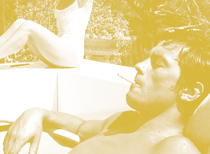 Man lounging poolside with a cigarette in his mouth while another person sits at the edge in a swimsuit, sunny scene behind them.