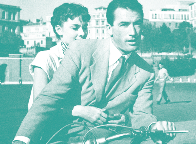 Young couple in vintage clothing ride a bicycle together; man in a suit at the handlebars with a woman clinging from behind in a park setting.