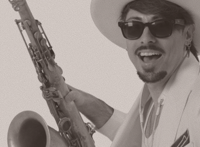 Man wearing sunglasses and a hat, playing a saxophone and smiling at the camera.
