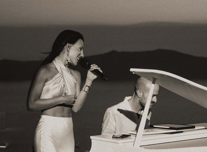 Woman singsinto a microphone beside a white grand piano, performing outdoors at sunset with hills in the background.