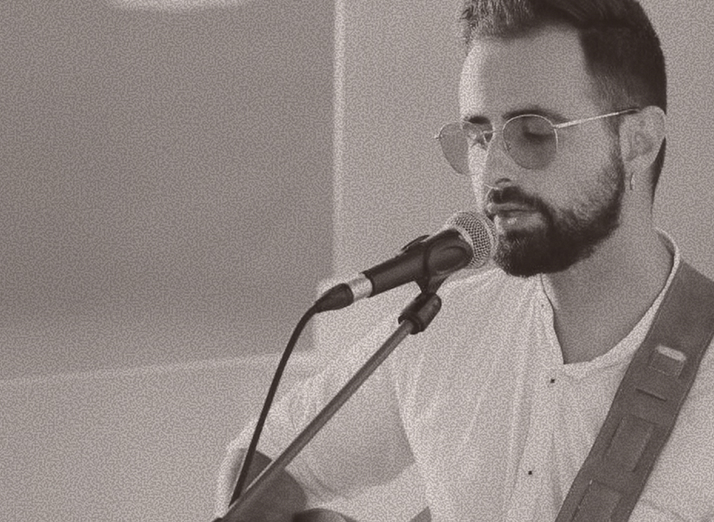 Male musician with a beard and sunglasses sings into a microphone while playing an acoustic guitar on stage, wearing a white shirt.